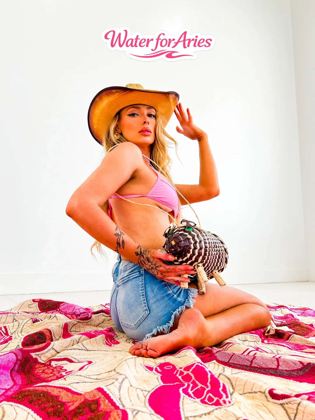A woman in a cowboy hat and pink bikini top poses with a necklace and denim shorts.