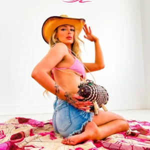 A woman in a pink bikini top and denim shorts poses with a cowboy hat and a bag.